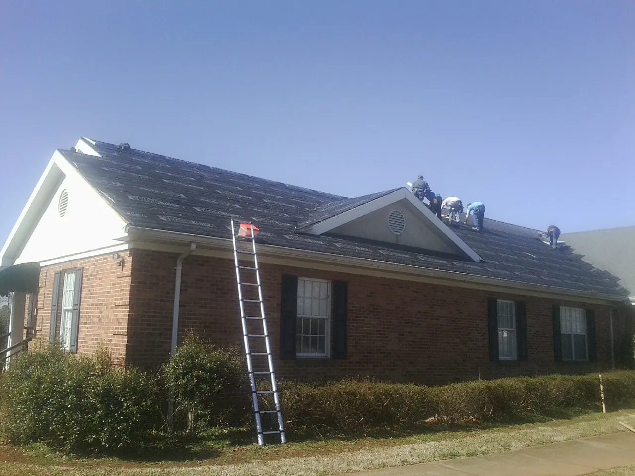 Crew tearing shingles off a brick ranch roof with a ladder set — Metal Roof Repair work by our crew in Berthoud