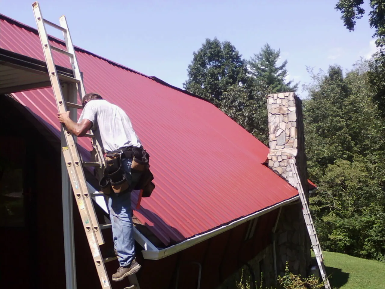 Roofer on a ladder beside a red metal roof and stone chimney — Soffit & Fascia Repair by Roofing Brothers in Berthoud